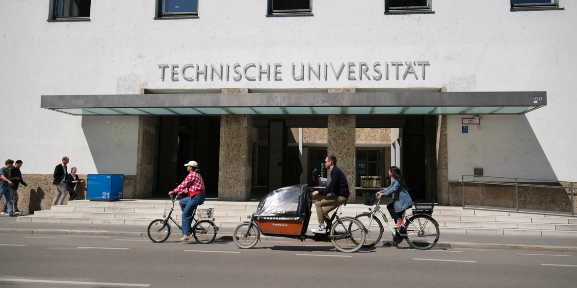 Three cyclists cycle in front of the TUM main entrance in Munich.